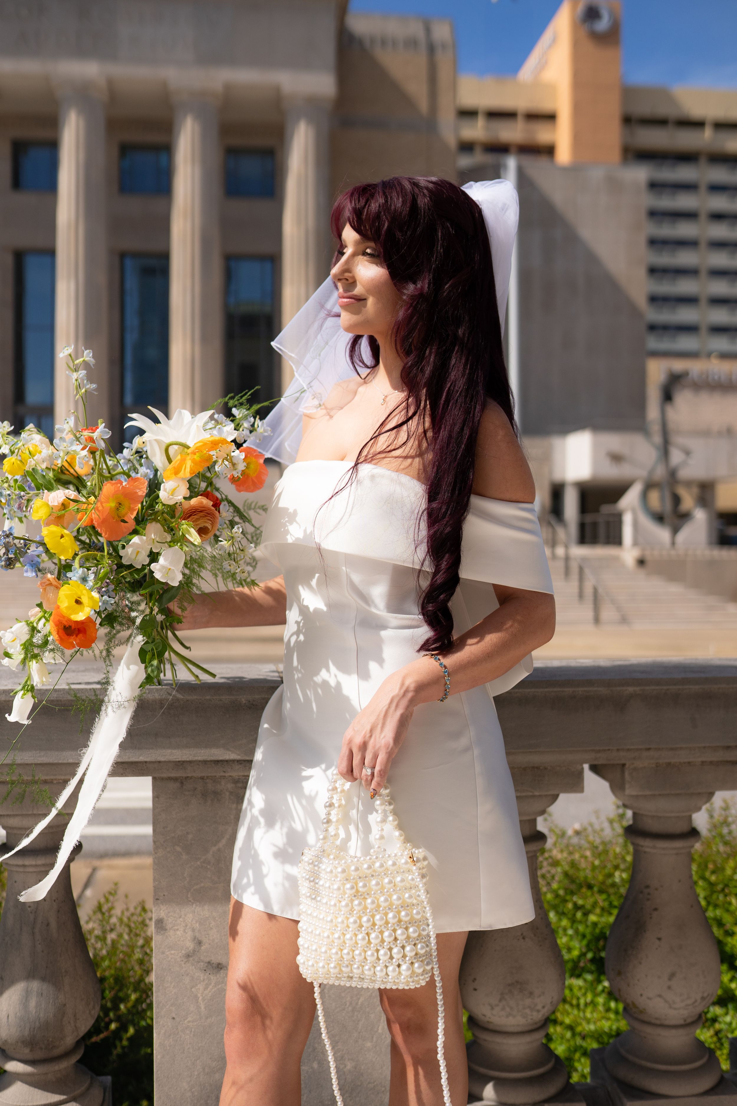 Bride in short white dress holding colorful bouquet poses against urban architectural backdrop.