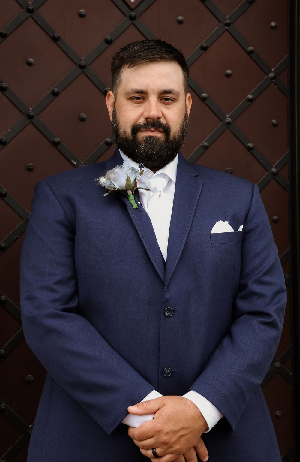 Handsome groom in navy blue suit with white boutonniere posing before ornate wooden door.