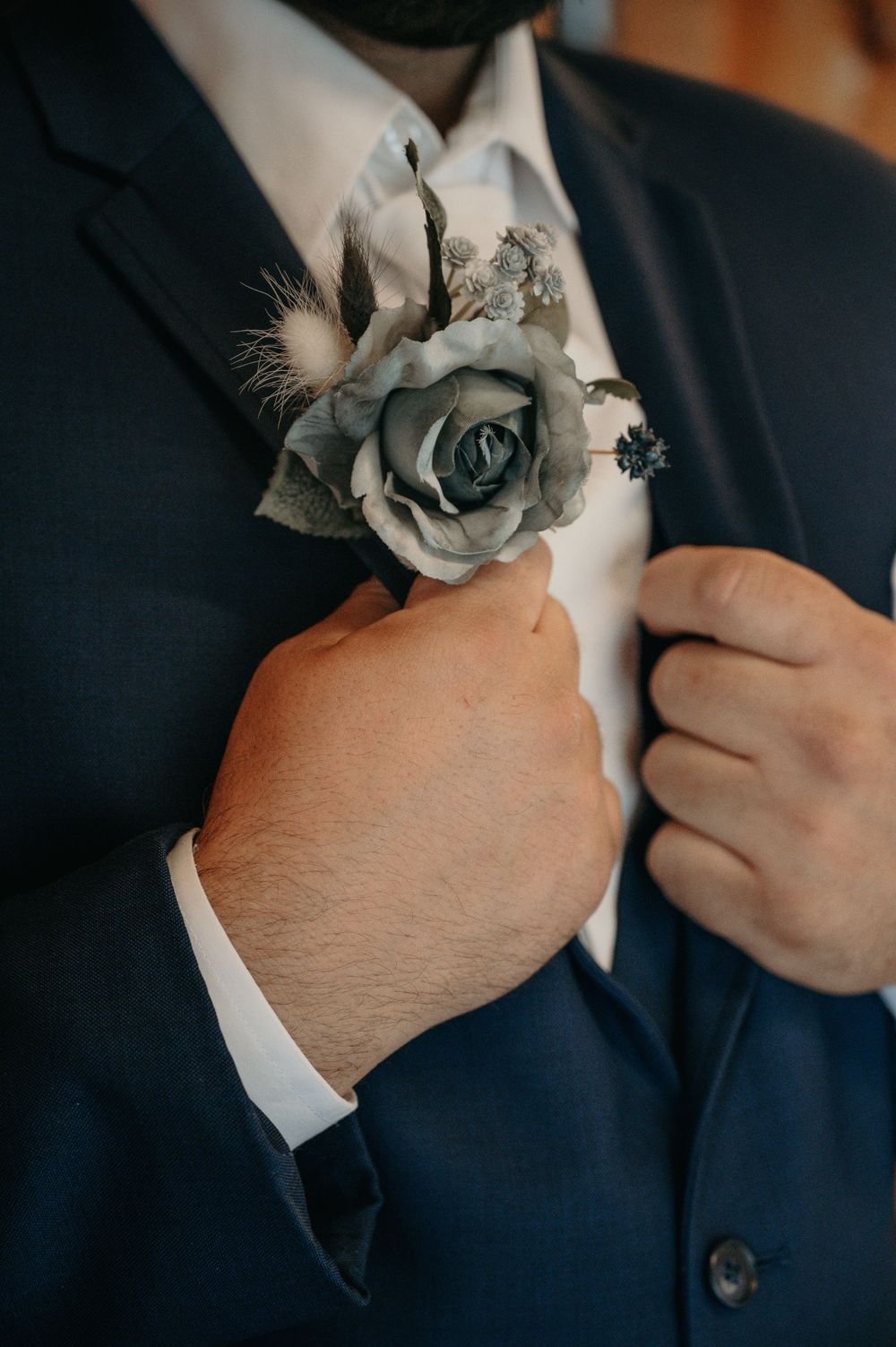 Groom in navy suit adjusts gray rose boutonniere with feather accent on lapel for wedding ceremony.