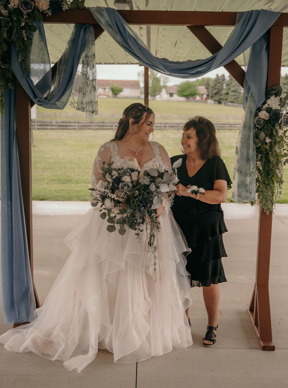 Bride in ruffled lace gown holding cascading bouquet stands with woman under blue draped floral arch.
