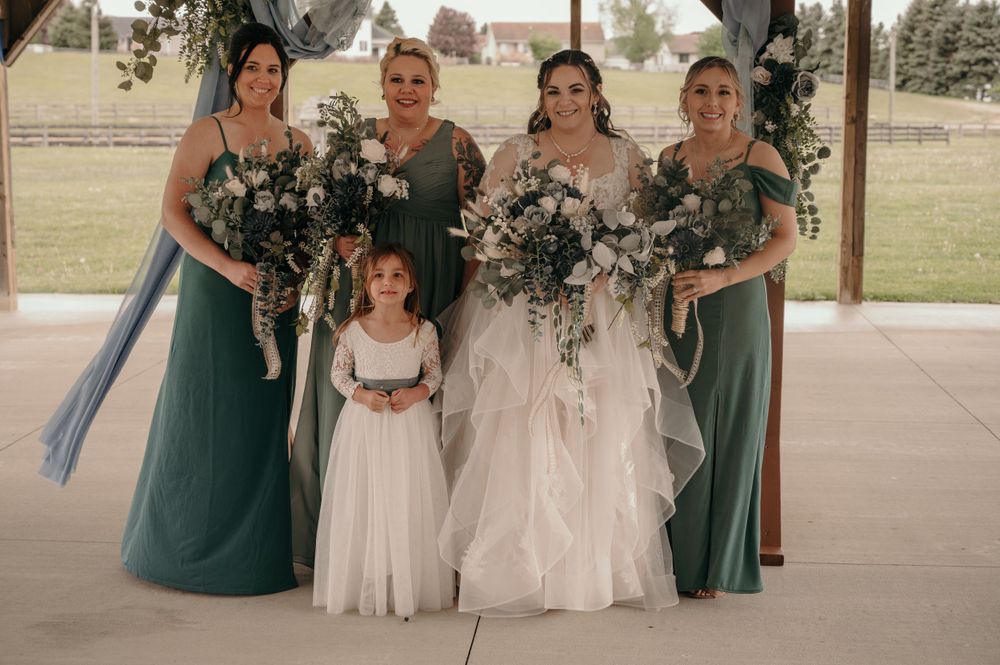 Bride with bridesmaids in teal gowns and flower girl in white pose under blue draped ceremony arch.