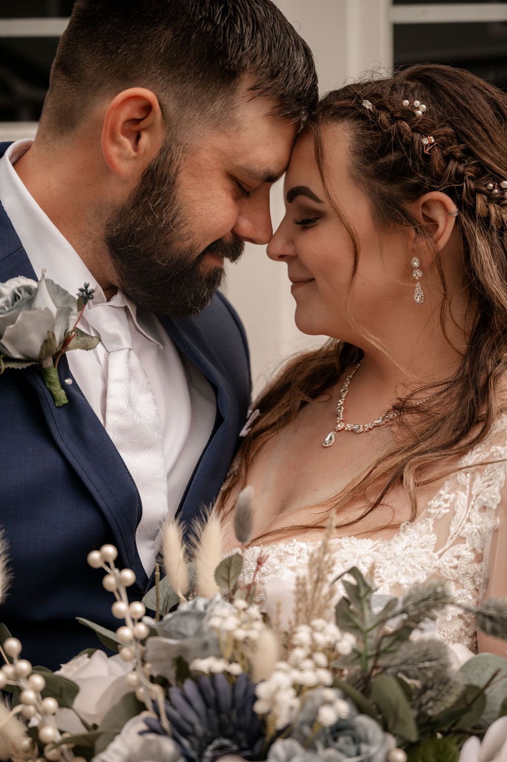 Bride and groom touching foreheads tenderly over a bouquet of dusty blue and white flowers.
