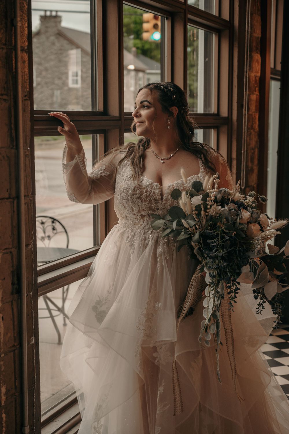 Bride in lace tulle gown holding bouquet gazing out a vintage window with soft natural light.