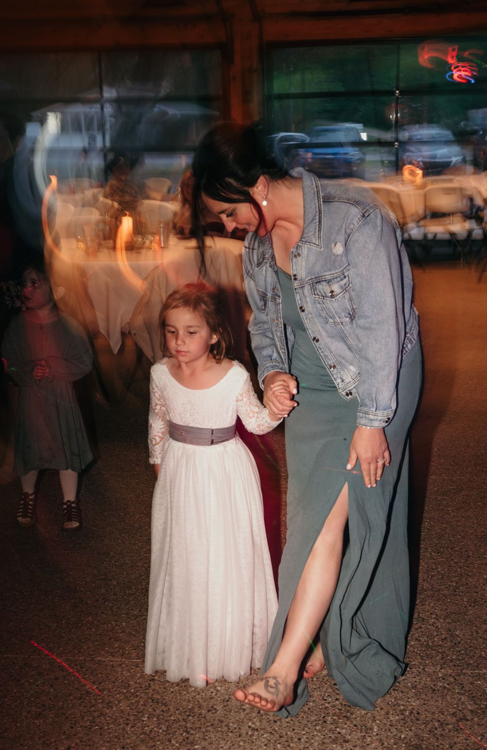 Woman in denim jacket and green dress dancing with young flower girl at wedding reception.