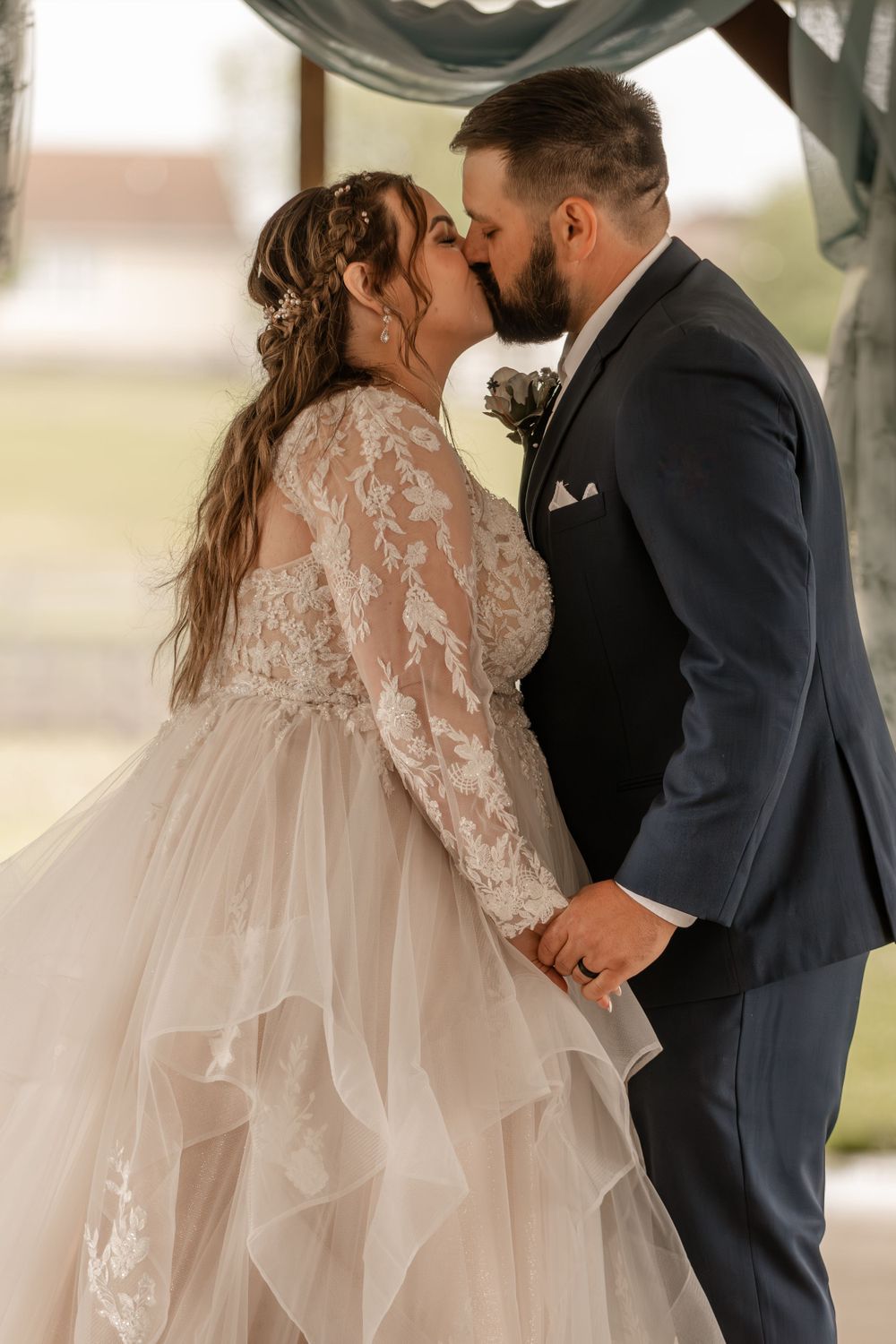 Bride and groom sharing first kiss at outdoor wedding ceremony under a rustic arch.