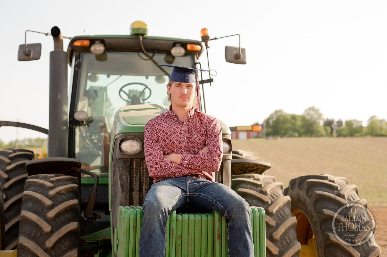 Graduate sits confidently on green tractor in agricultural field.