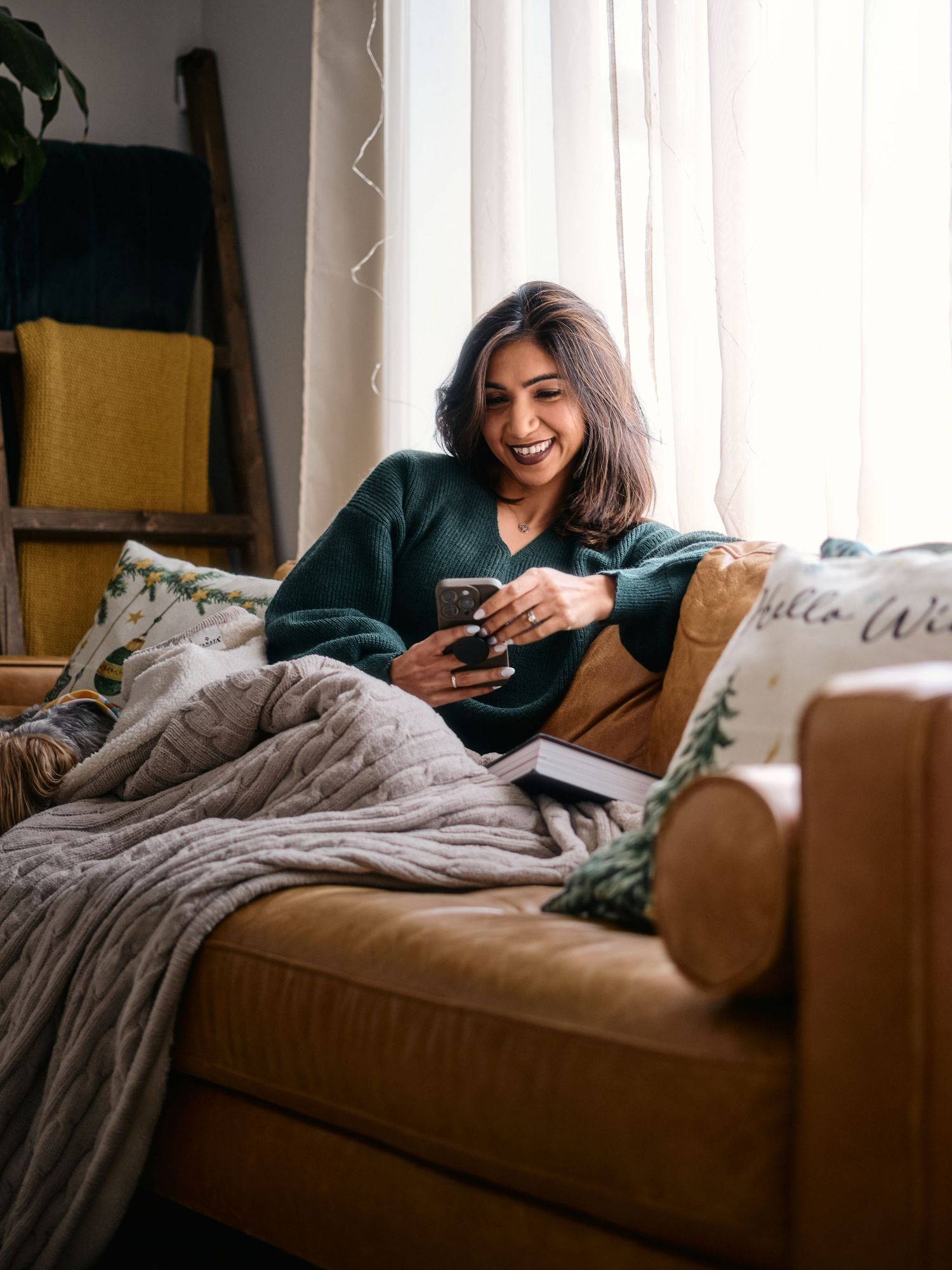 Editorial portrait of a model relaxing on a cozy tan couch with a smartphone, wearing a dark green sweater and covered with a gray blanket.