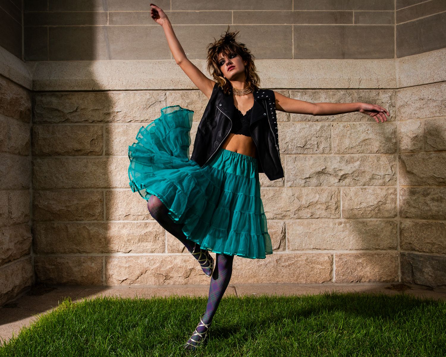 Dynamic fashion pose with teal tulle skirt against stone wall and grass.