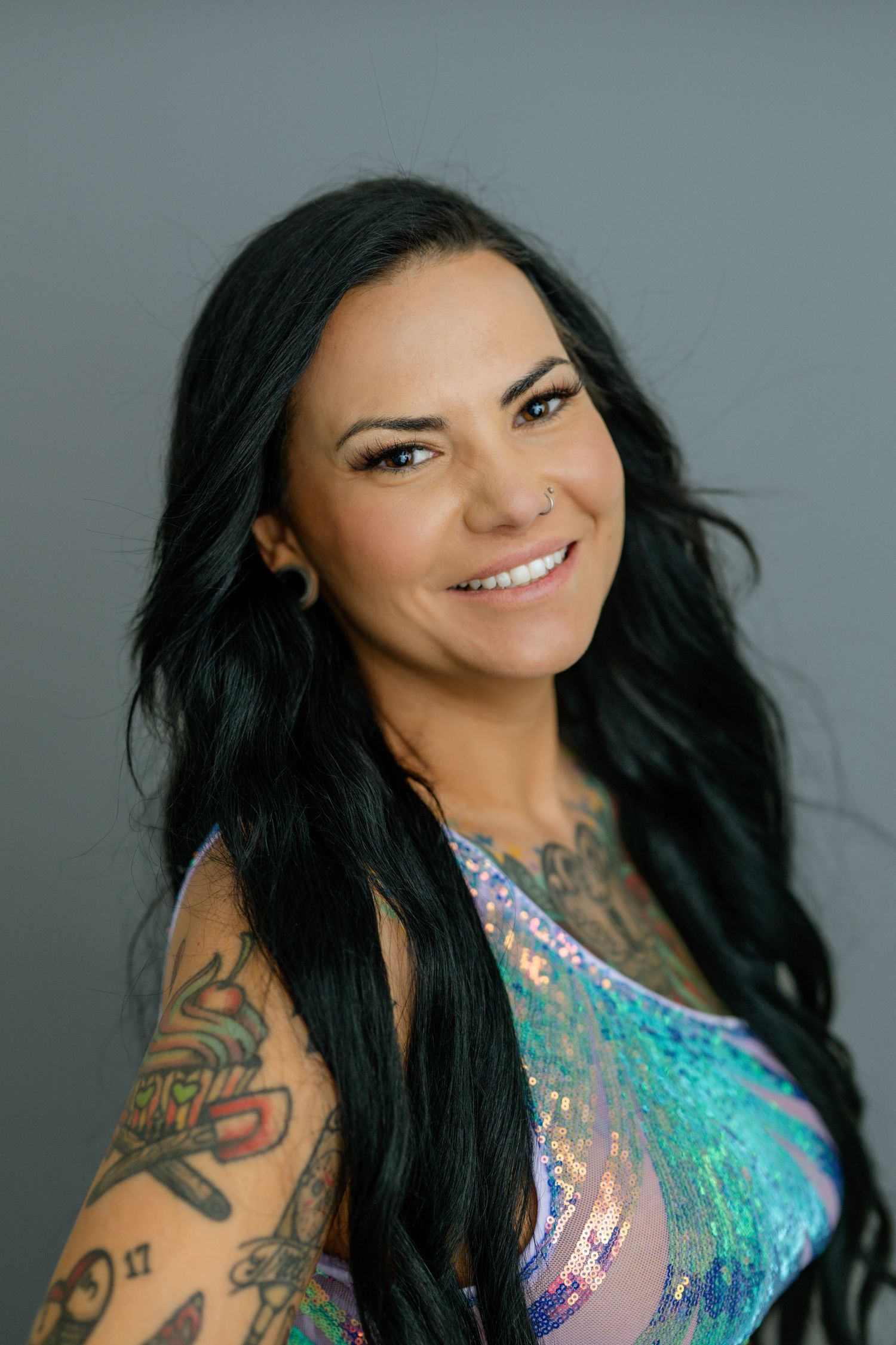 Woman with tattoos and dark hair wearing iridescent top smiles against grey background.