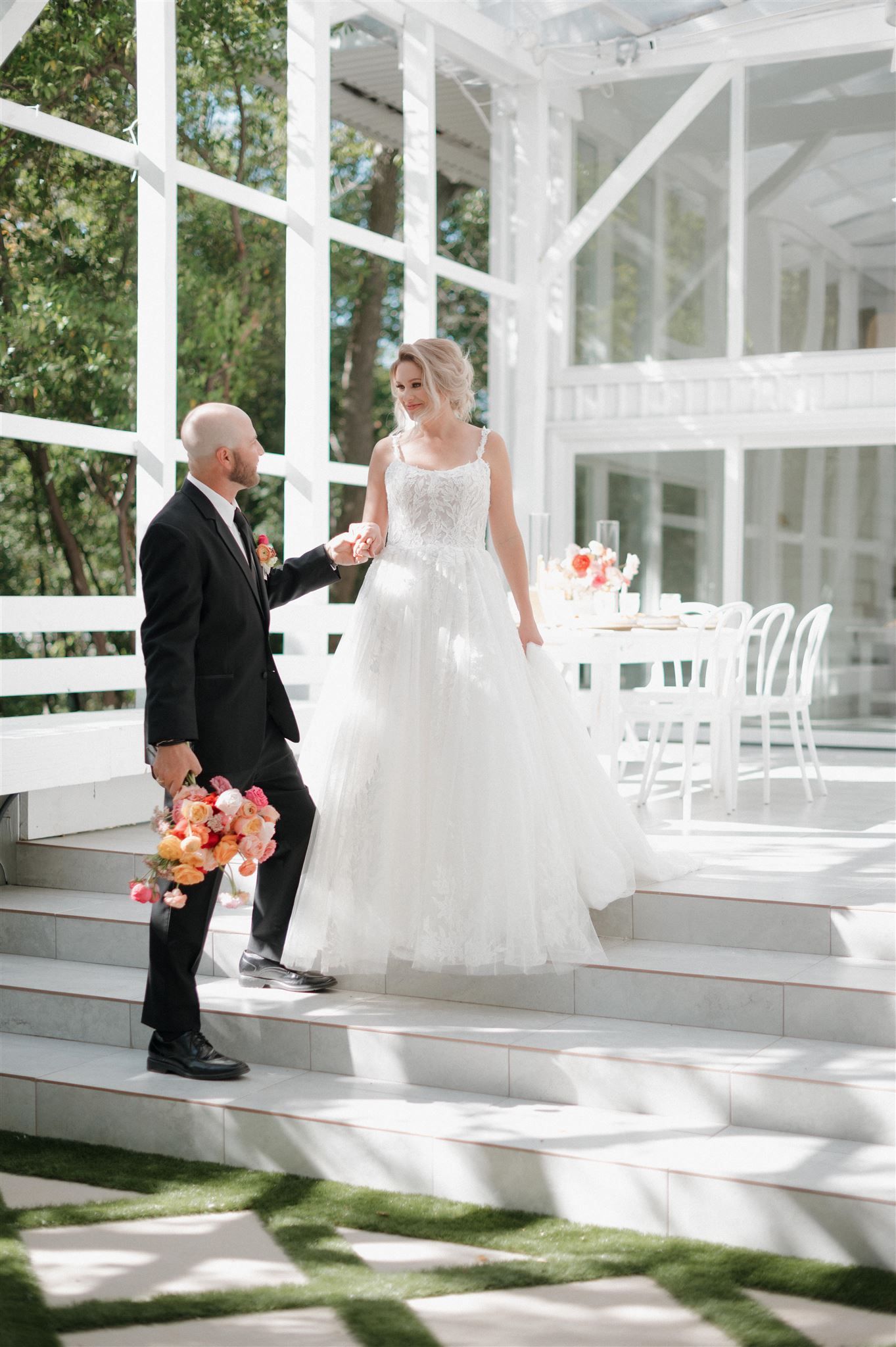A couple in wedding attire stand on white steps of an elegant gazebo surrounded by greenery and white decor.