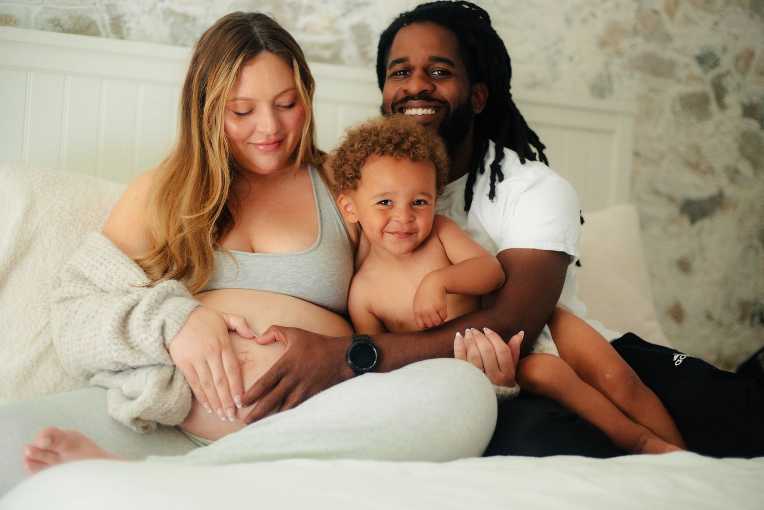 husband and wife with toddler son snuggled in bed making a heart shape with their hands on moms pregnant belly during an in home session