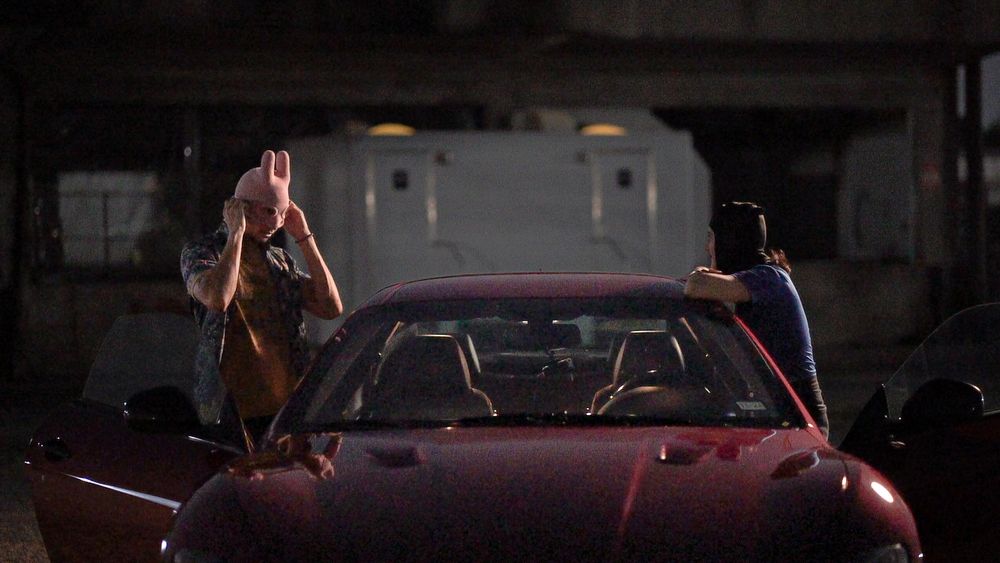 Two people interacting near maroon car in dimly lit parking area.
