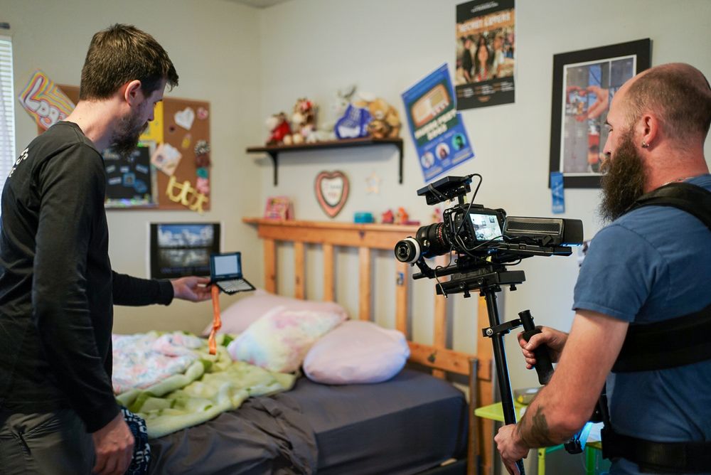 Two camera operators filming in a bedroom with toys and posters visible on the film set.