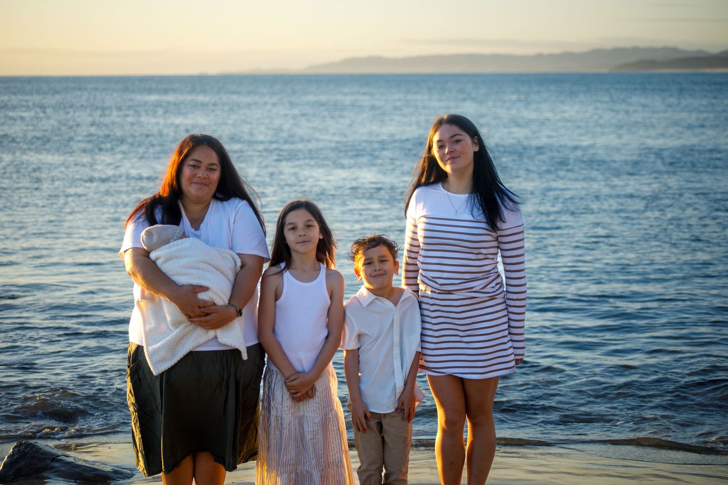Family of four with baby posing on waterfront dock at golden sunset hour.