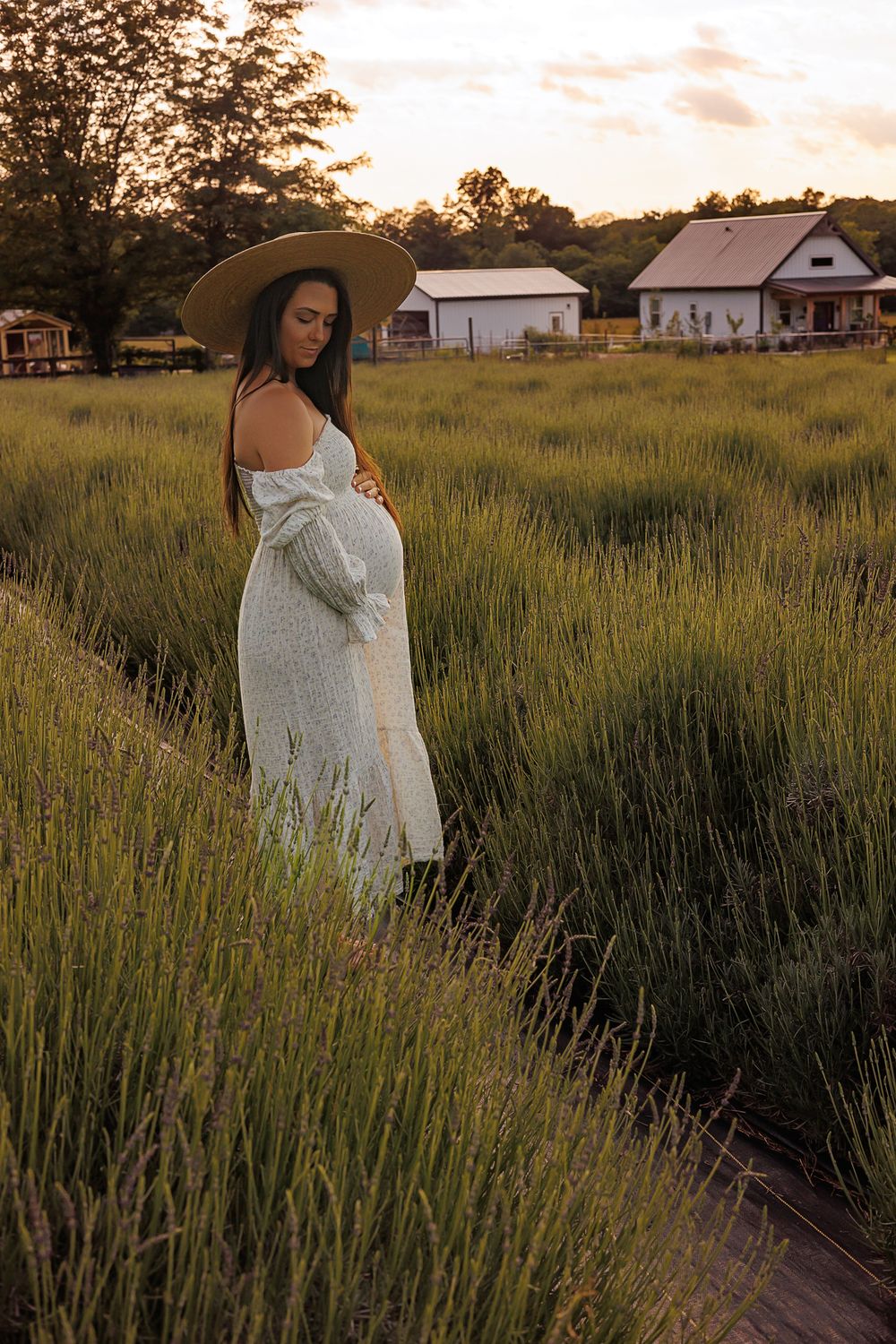 Silhouette of expecting mother in white dress with farmhouse in background at sunset.