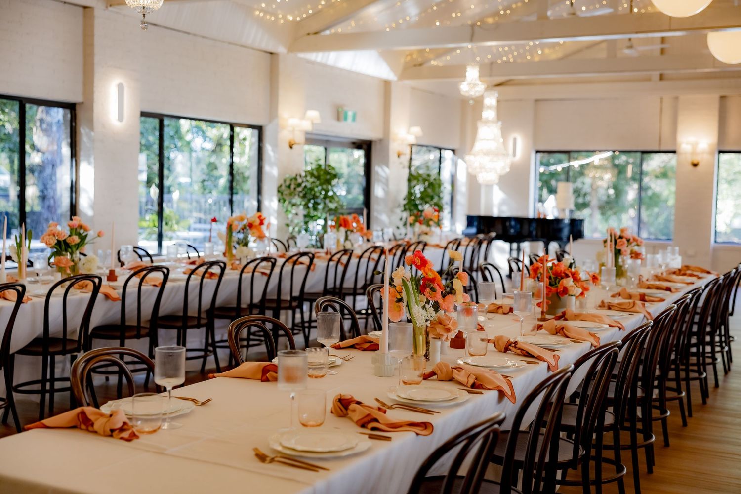 Long reception table with bentwood chairs and orange floral centerpieces in bright dining room.