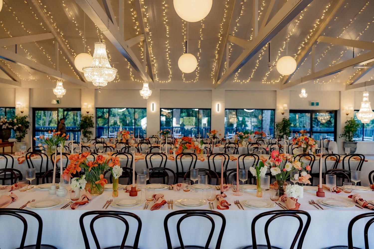 Wedding venue with string lights, chandeliers and bentwood chairs set for reception dinner.