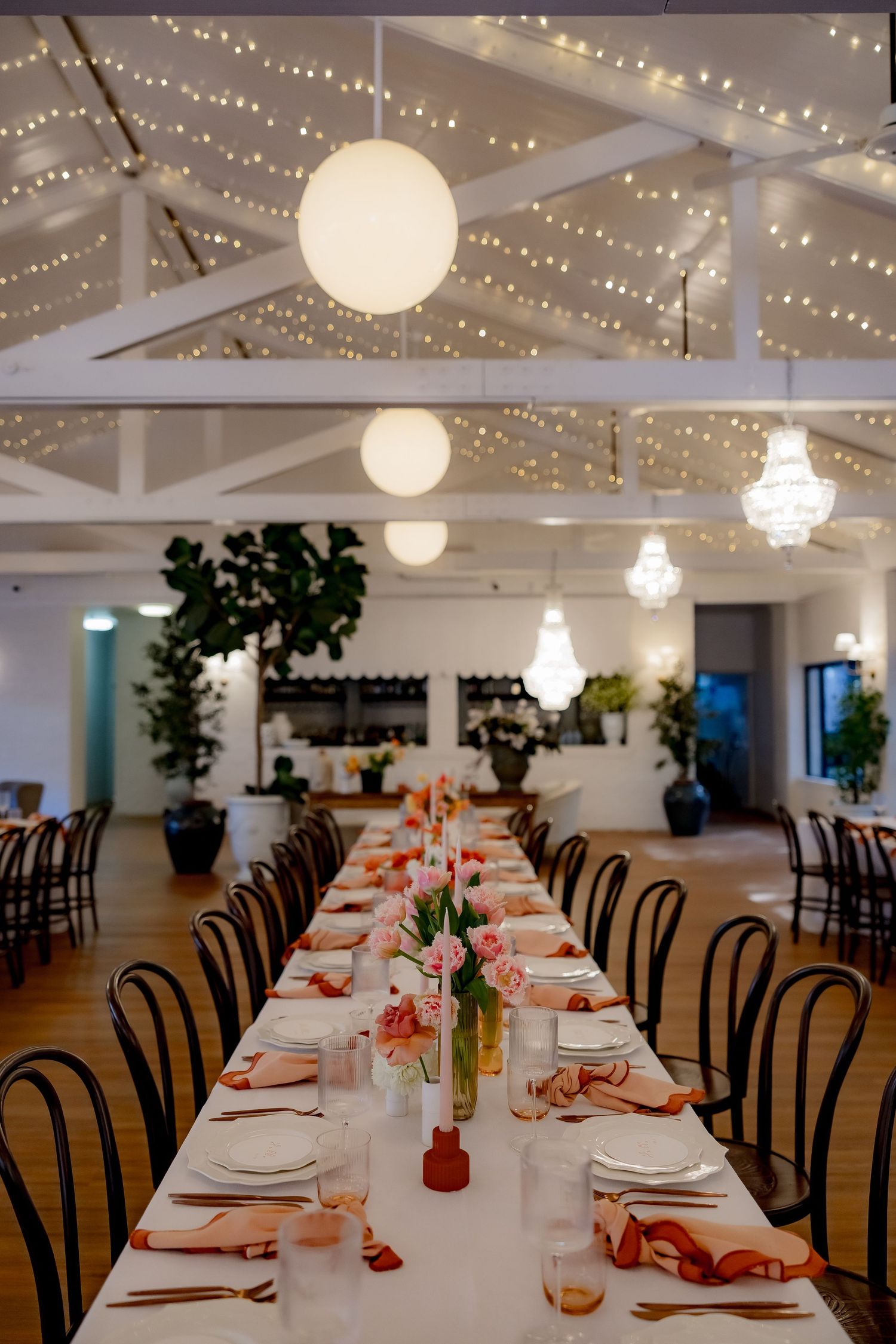 Long banquet table set under twinkling lights and globe pendants in modern white venue.