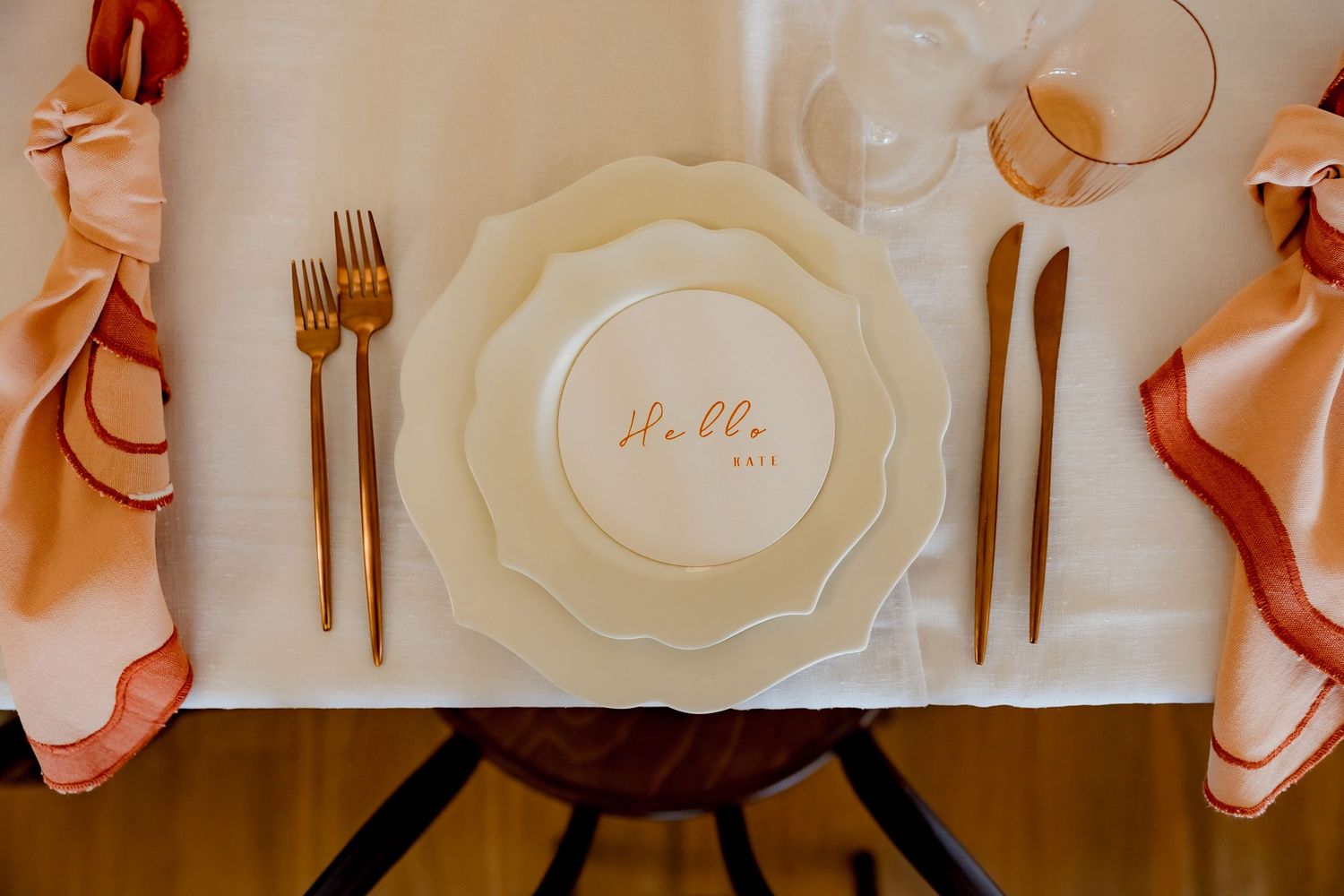 Elegant place setting with gold flatware, cream-colored plate, and peach napkin on white tablecloth.