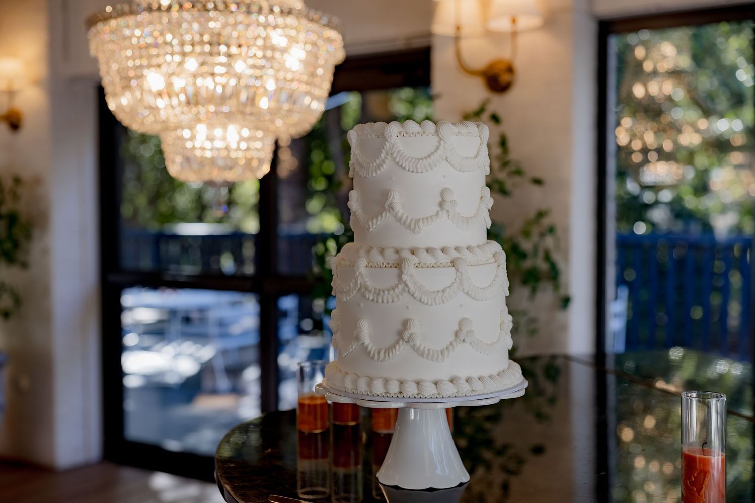 Elegant white three-tiered wedding cake displayed under crystal chandelier lighting.