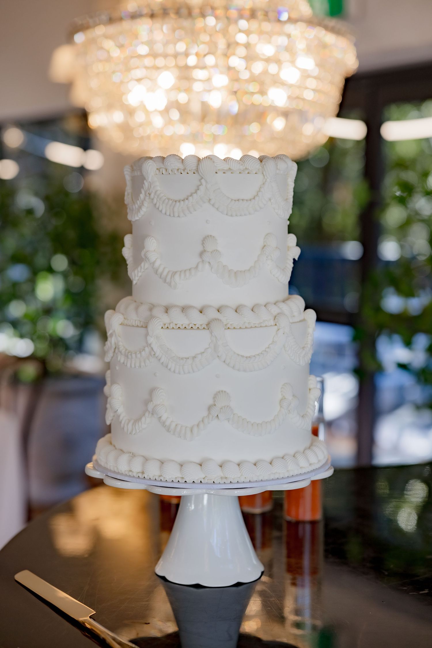 Classic white wedding cake with delicate scalloped piping details under a crystal chandelier.