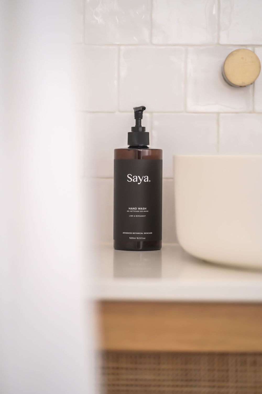 A minimalist brown Saya soap dispenser sits on a bathroom counter against white tile.