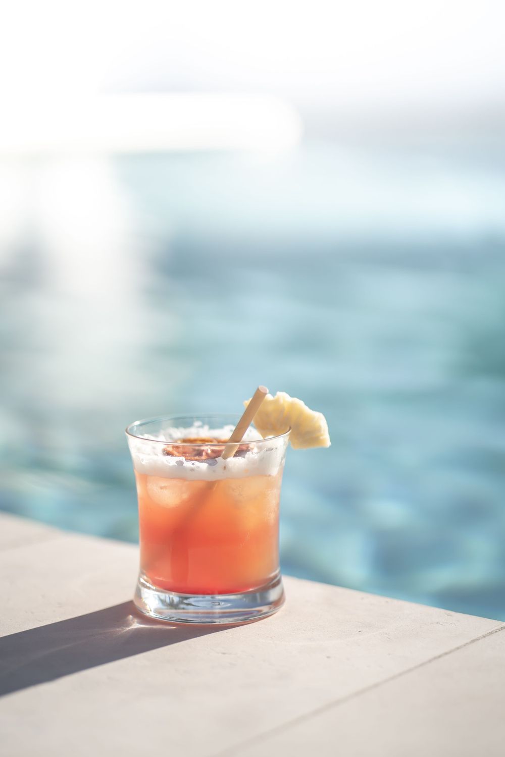 Refreshing pink cocktail with pineapple garnish overlooking ocean waters on a sunny day.
