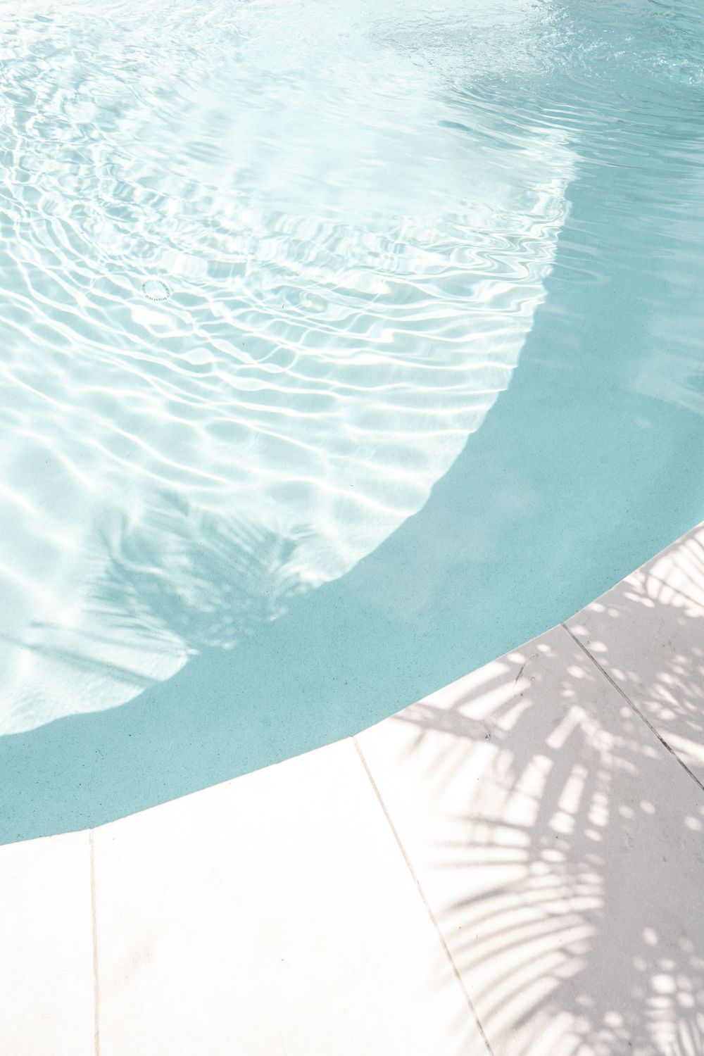 Crystal clear swimming pool water with ripples and palm tree shadows on white pool deck.