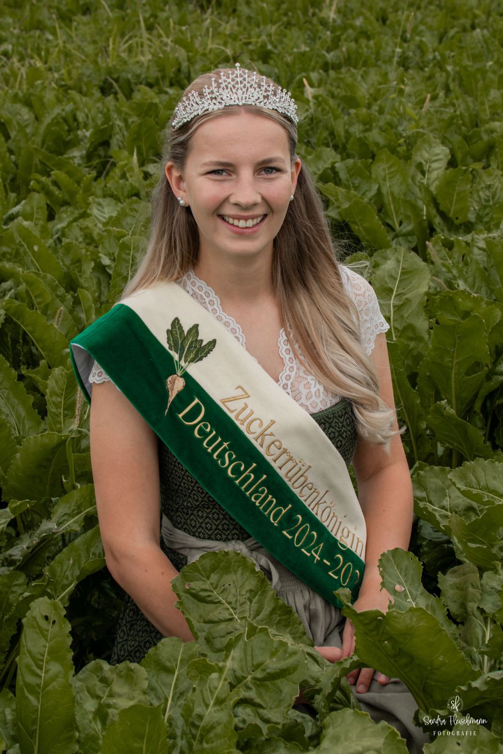A pageant winner wearing a crown and green sash poses in a leafy field.
