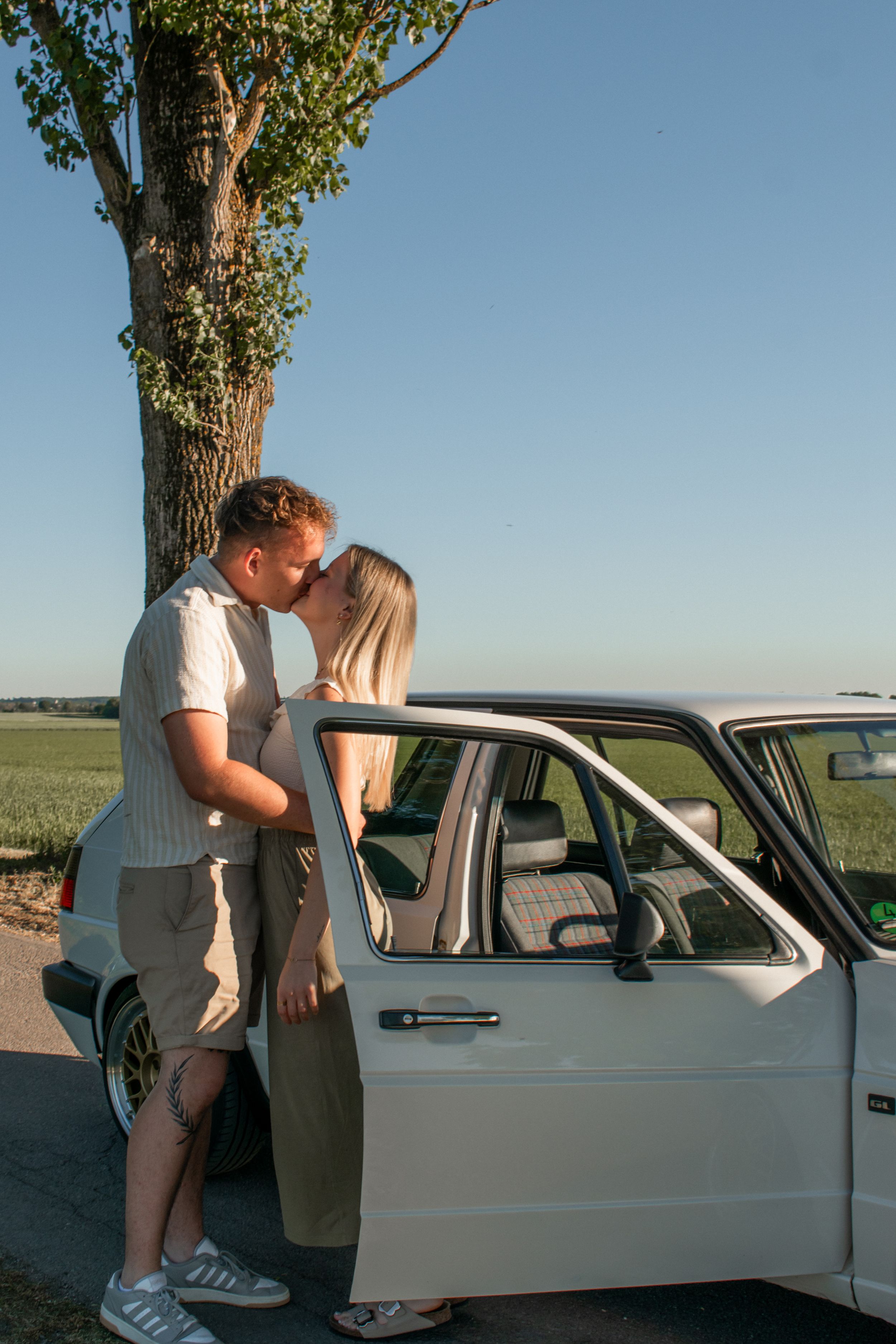 A couple sharing a romantic kiss beside a vintage white car near a tree at sunset.