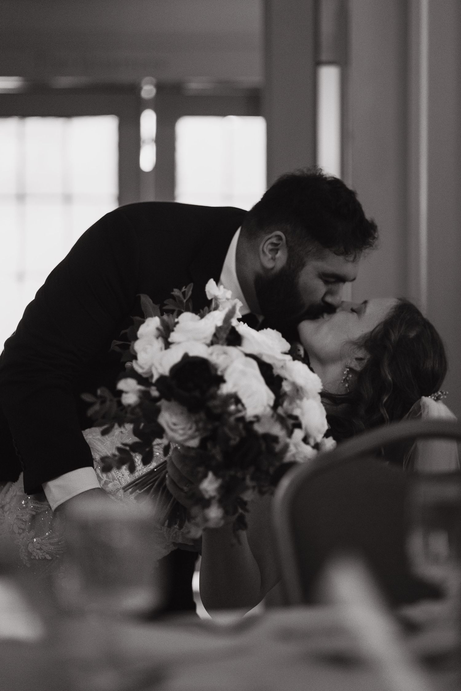 A romantic black and white wedding moment as a couple shares a kiss while holding a beautiful floral bouquet at their reception.