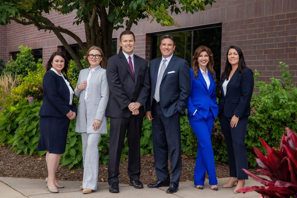 Business team of six people in suits posing outside a brick building with landscaping.