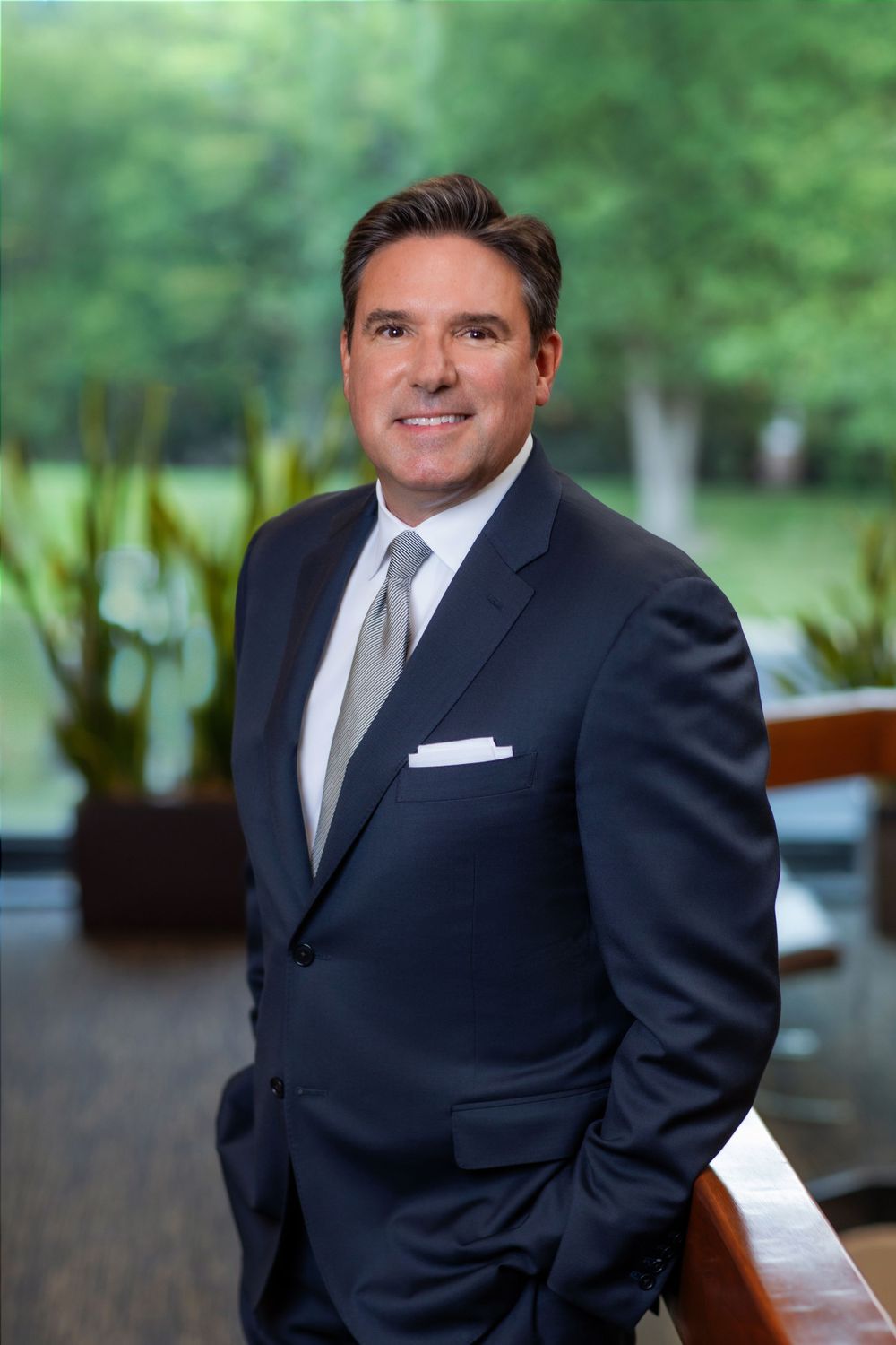 Professional headshot of executive in dark suit against blurred outdoor background.