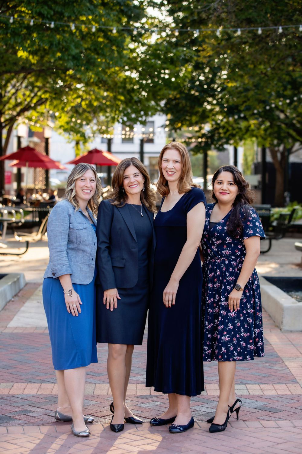 Four professionals in business attire pose together on brick walkway with trees and cafe setting.