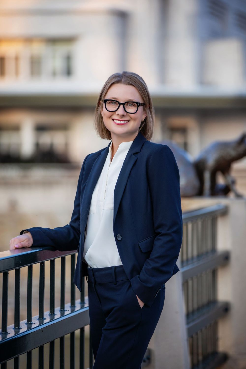 Professional attorney wearing glasses and navy suit poses against railing with urban background.
