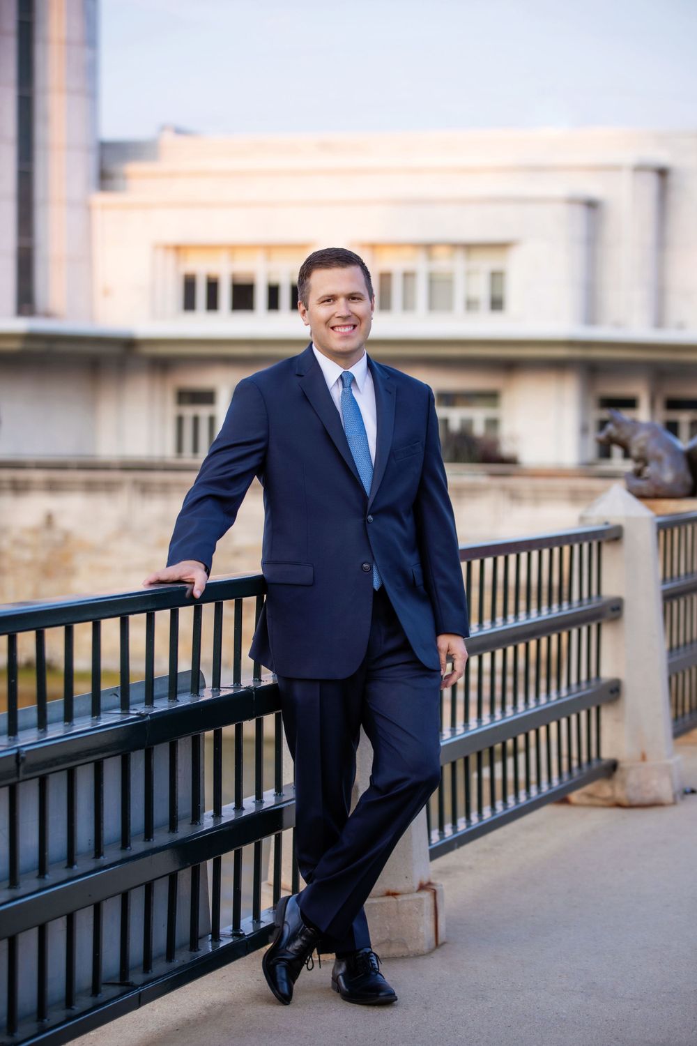 Professional attorney in navy suit and blue tie poses confidently against outdoor railing.