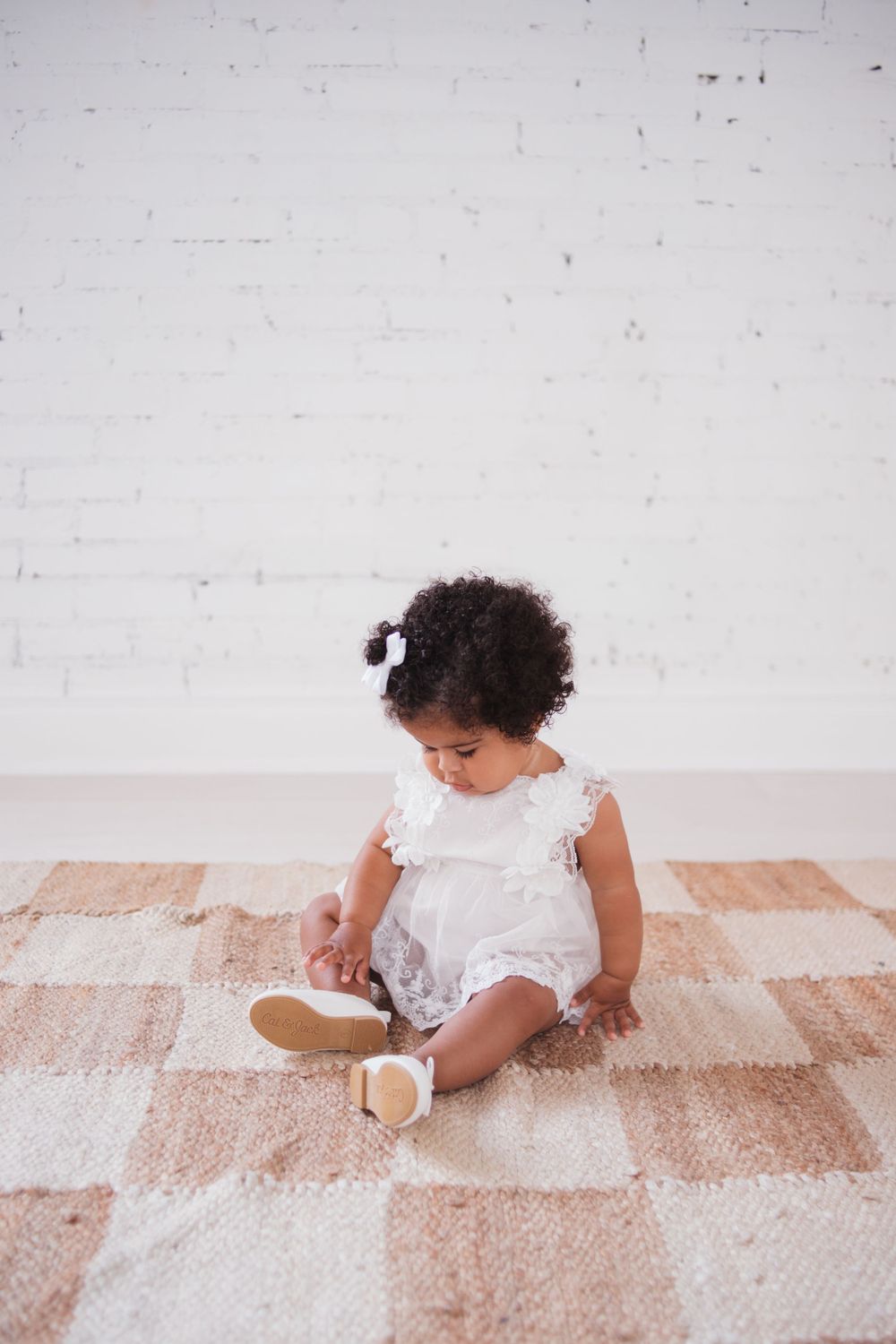 Small child in white lace dress sitting on checkered rug indoors.