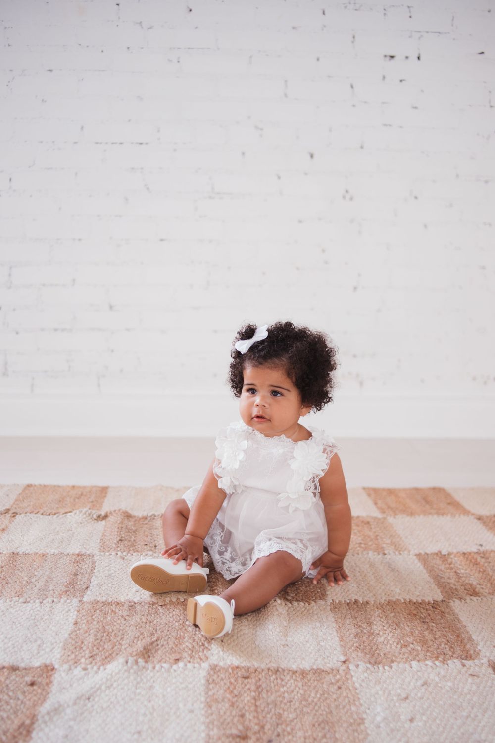 Baby in white lace outfit sits on checkered rug against white wall.