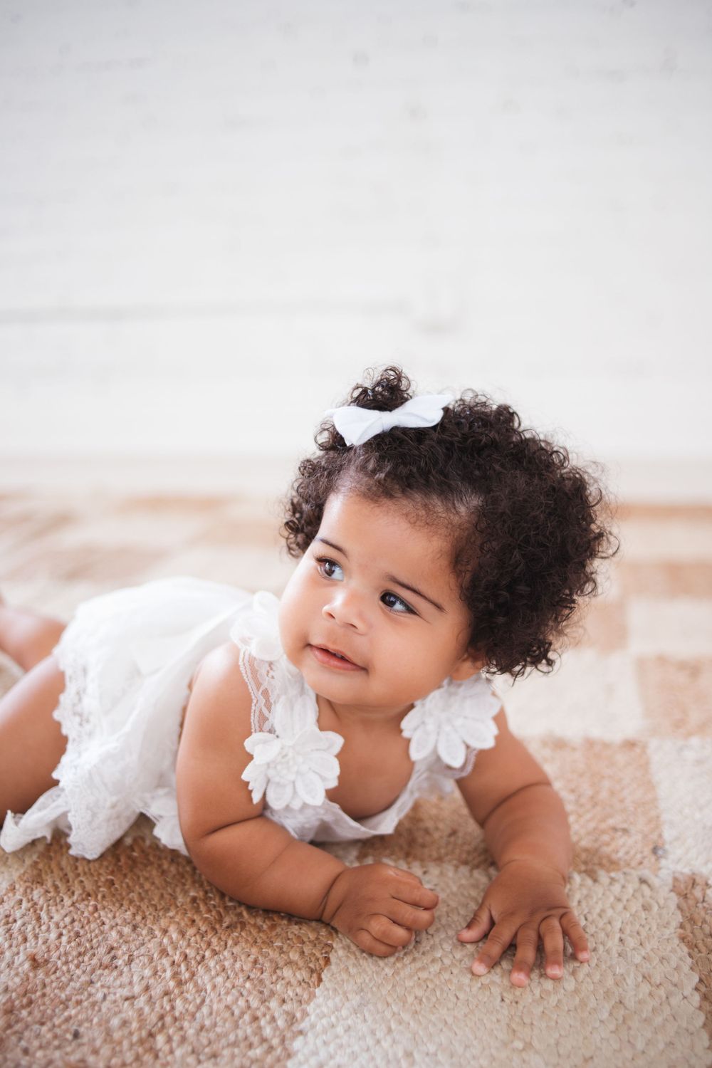 Baby wearing white lace dress lying on beige carpet.