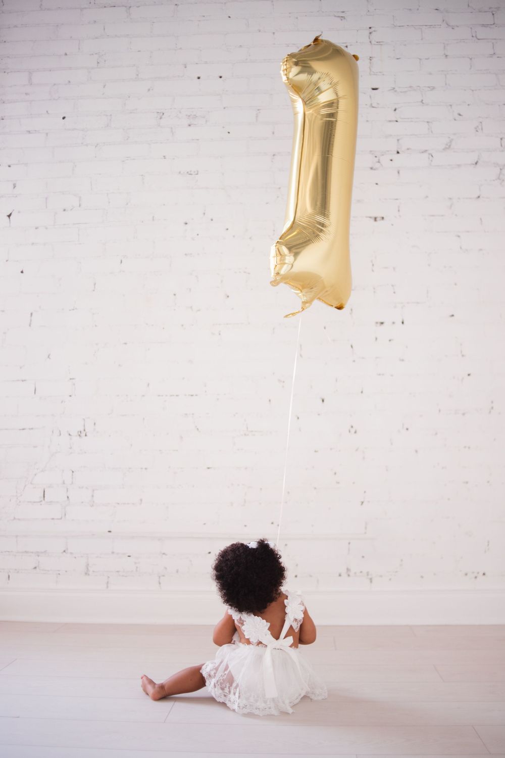Baby in white dress sits beneath large gold number one balloon against white wall.