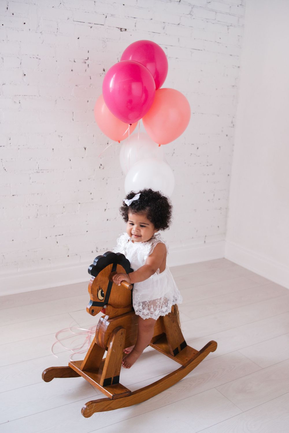 Baby rides wooden rocking horse with pink and white balloons floating above.