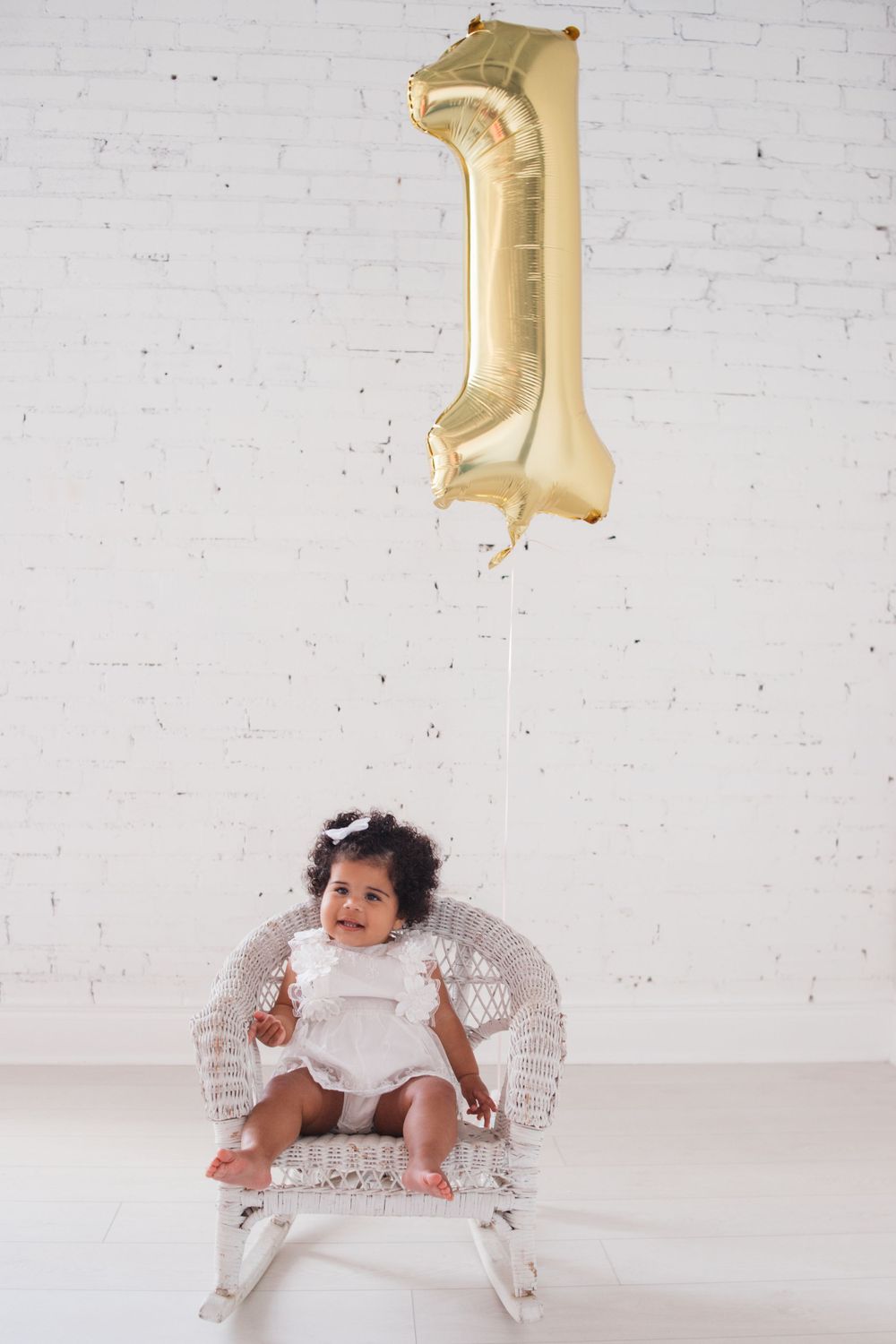 Baby sits in ornate chair under gold number one balloon against white wall.