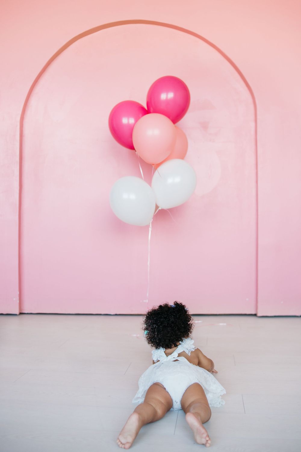 Baby in white dress lying near pink and white balloons against pink arch.