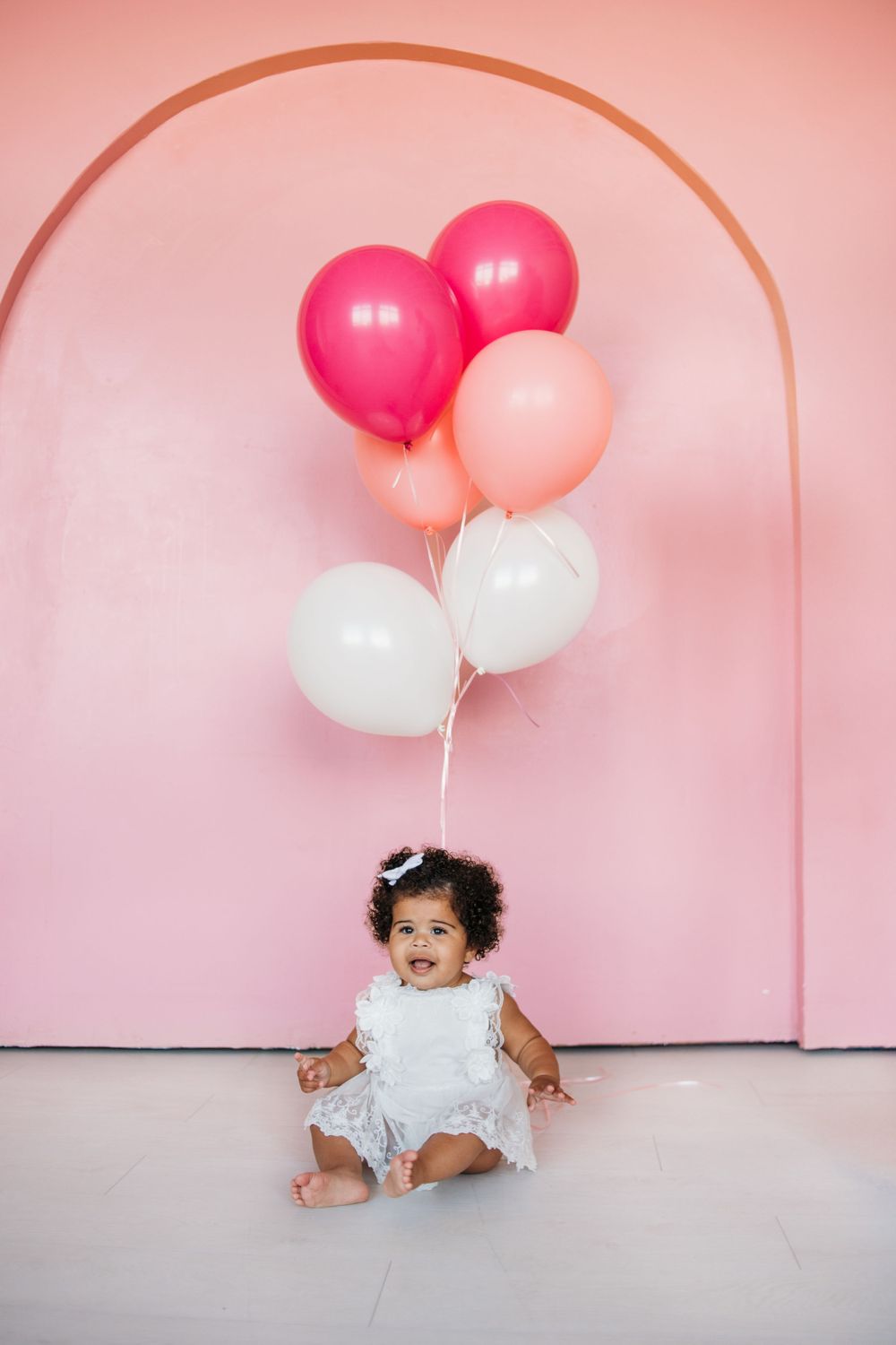 Baby sits beneath pink and white balloons against curved pink wall.