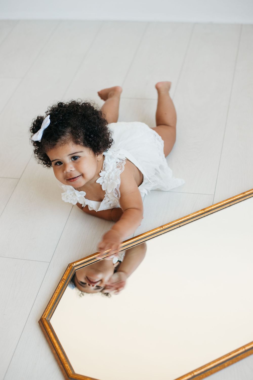 Toddler in white dress lying on floor next to decorative mirror.