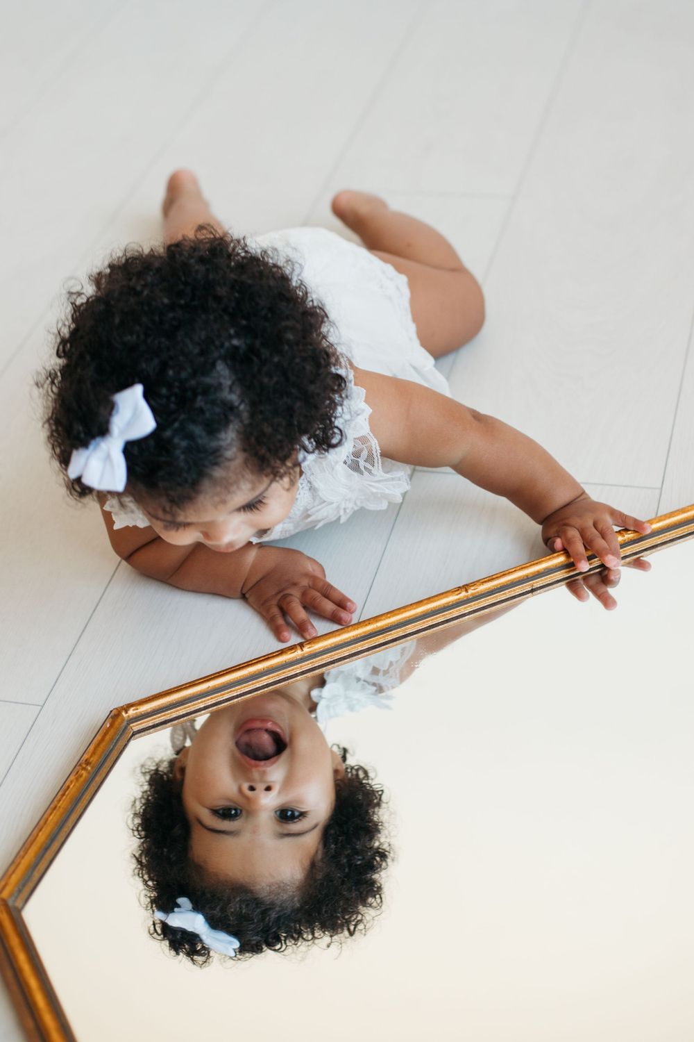 Baby in white dress looking at reflection in gold-framed mirror on floor.