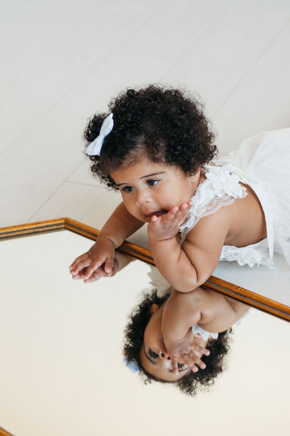 Child in white dress looking at reflection in gold-framed mirror on floor.