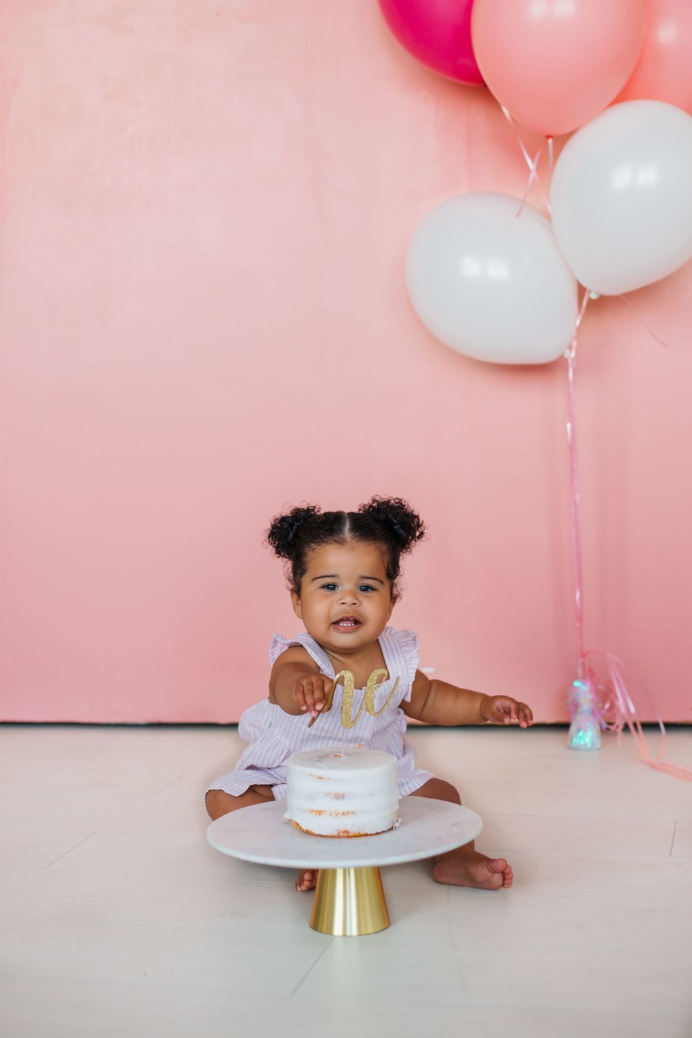 Baby in white dress with cake on gold stand against pink wall with balloons.