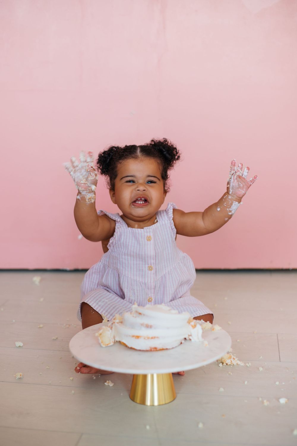 A toddler in a white dress raises frosting-covered hands while sitting at a cake smash photo session against a pink backdrop.
