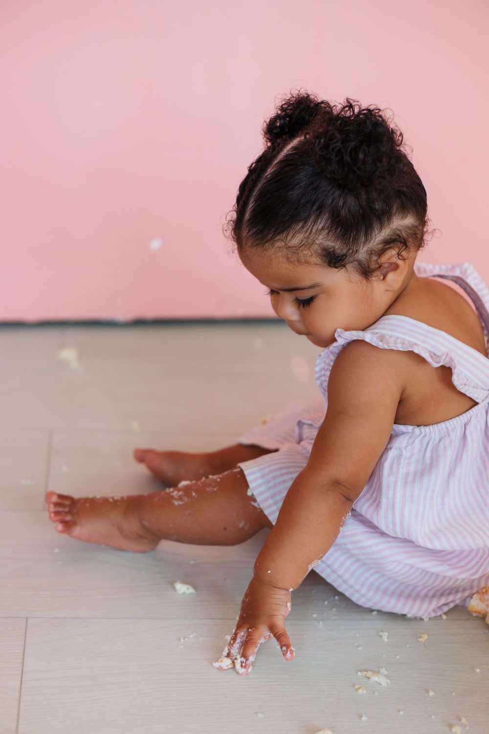 A small child in a white dress plays with cake frosting during a photoshoot on a gray floor.
